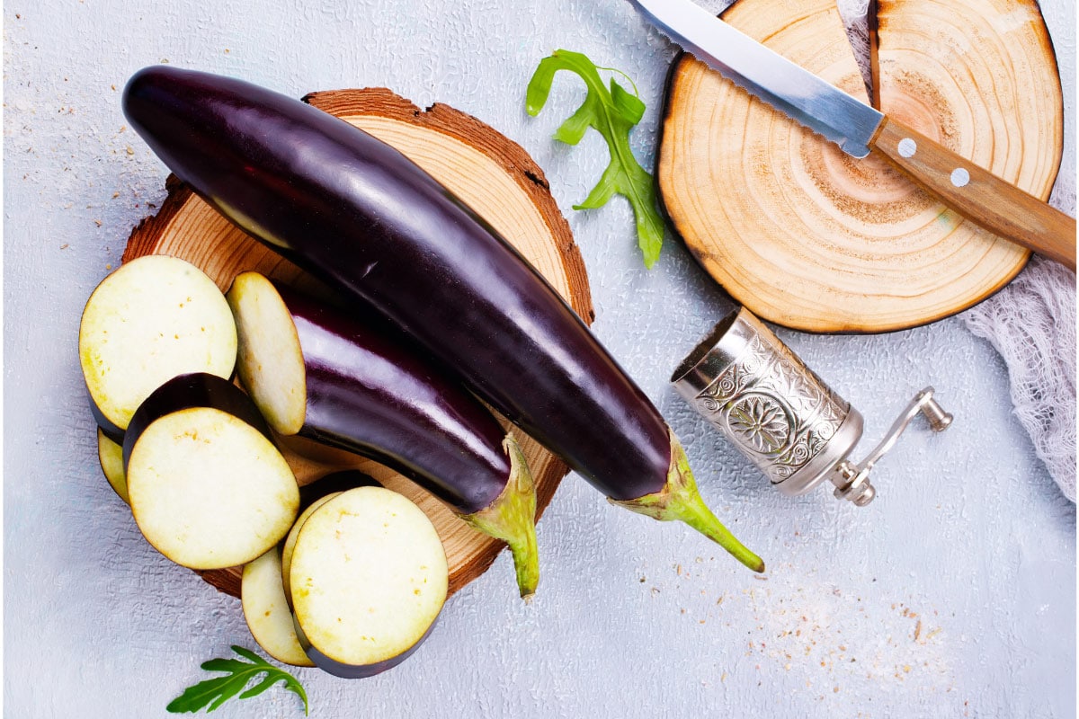 Diamond Eggplants are on the wooden stand. Next to it there some greens, knife and wooden stand | Hurry The Food Up