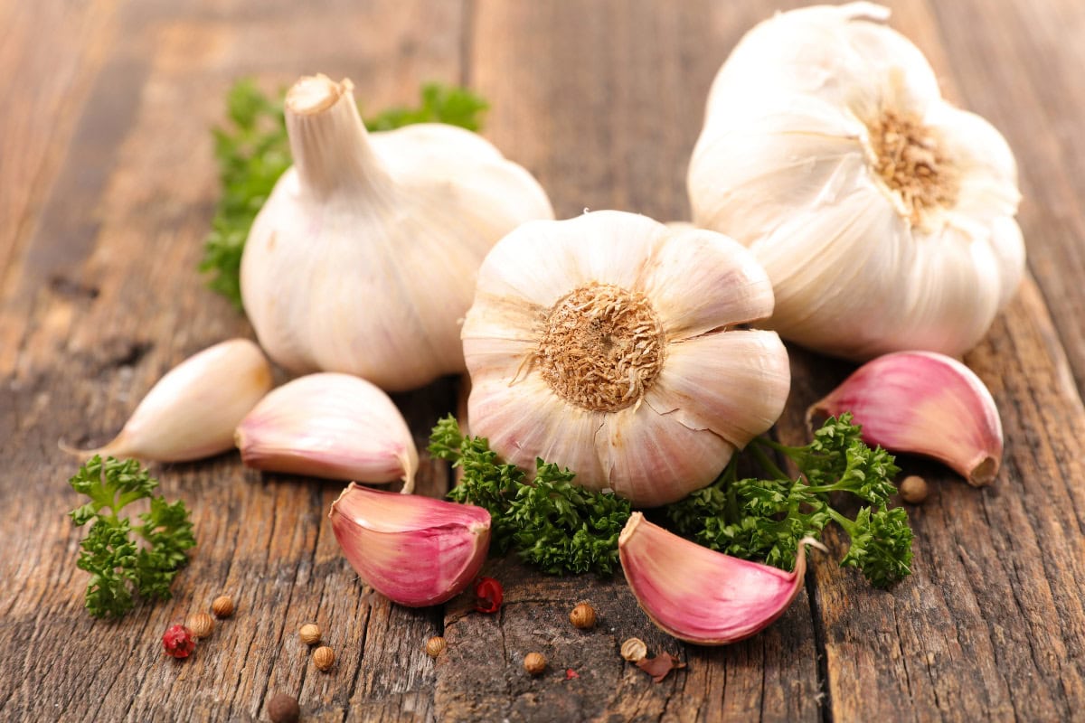 Three heads of dubna garlic are on the wooden surface. Next to them there're some garlic cloves | Hurry The Food Up