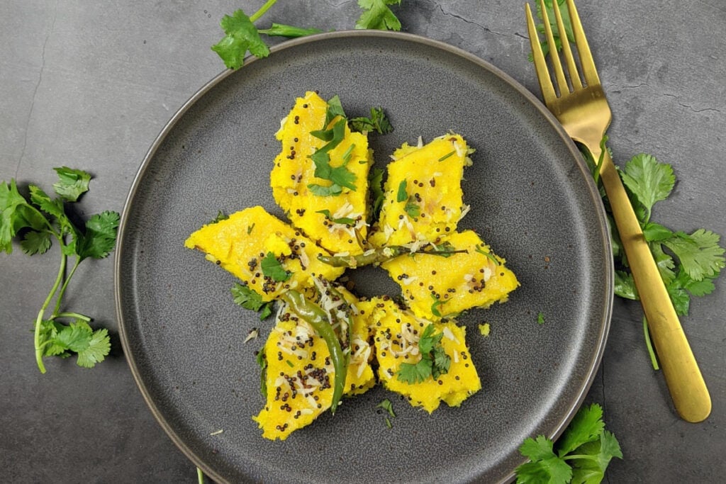 The dark plate with Dhokla. Near it there are some greens and bronze fork | Hurry The Food Up