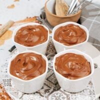 Four small white bowls with chocolate protein pudding stand on the plate. Behind them, there is a small wooden bowl with other cutlery | Hurry The Food Up