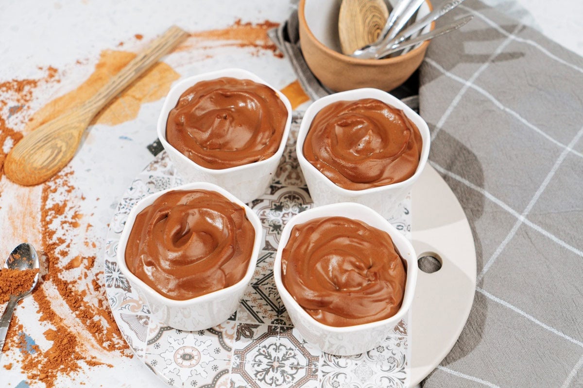 Four small white bowls with chocolate protein pudding stand on the plate. Near them, there is some cutlery | Hurry The Food Up