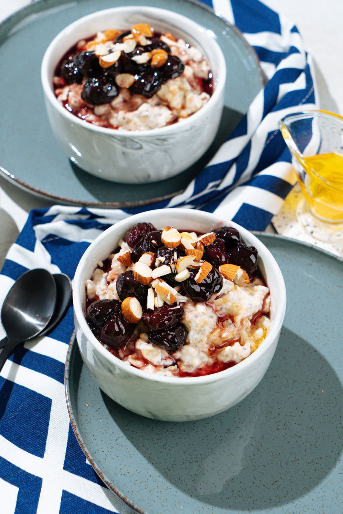 Two white bowls with protein oatmeal stand on the plates. Around them, there are two small spoons, glassware with honey, a plate with almonds, colorful napkin | Hurry The Food Up