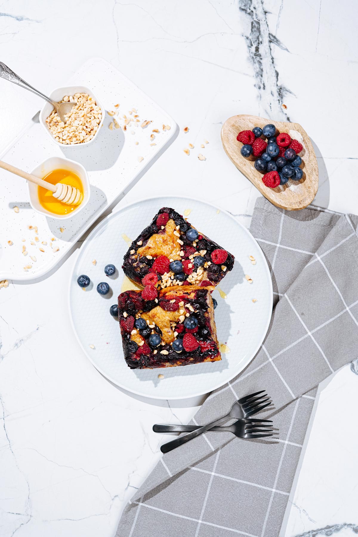 Bird's-eye view of a white plate with French toast is on a light surface. Next to it, there are grey napkin, two forks, a white tray with honey and chopped nuts, and a wooden plate with berries | Hurry The Food Up