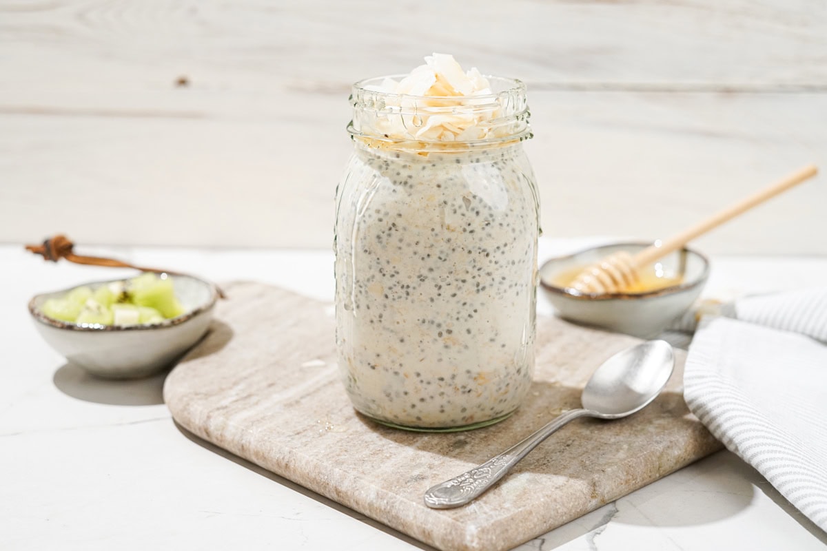 The jar with high-protein overnight oats and the spoon are on the marble сut board. Near it, there are two small bowls with kiwi and honey | Hurry The Food Up
