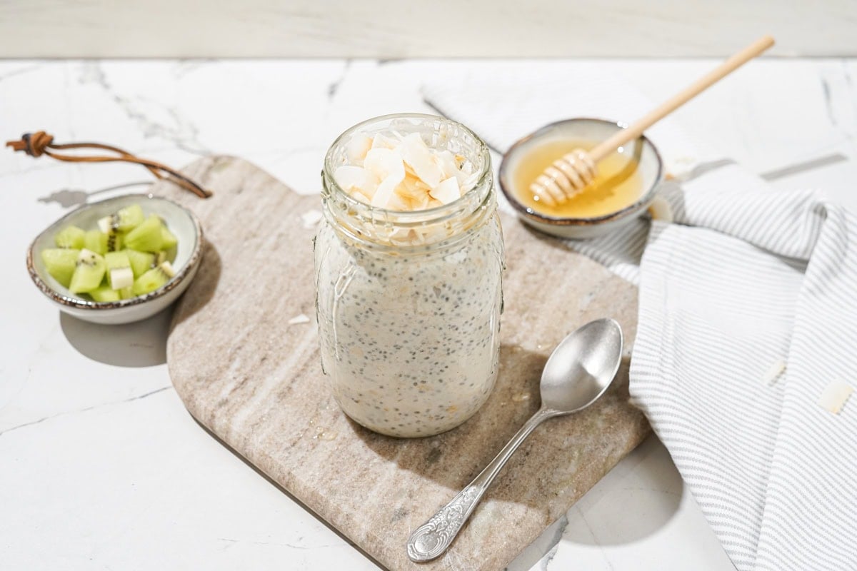 The jar with high-protein overnight oats and the spoon are on the marble сut board. Near it, there are two small bowls with kiwi and honey | Hurry The Food Up