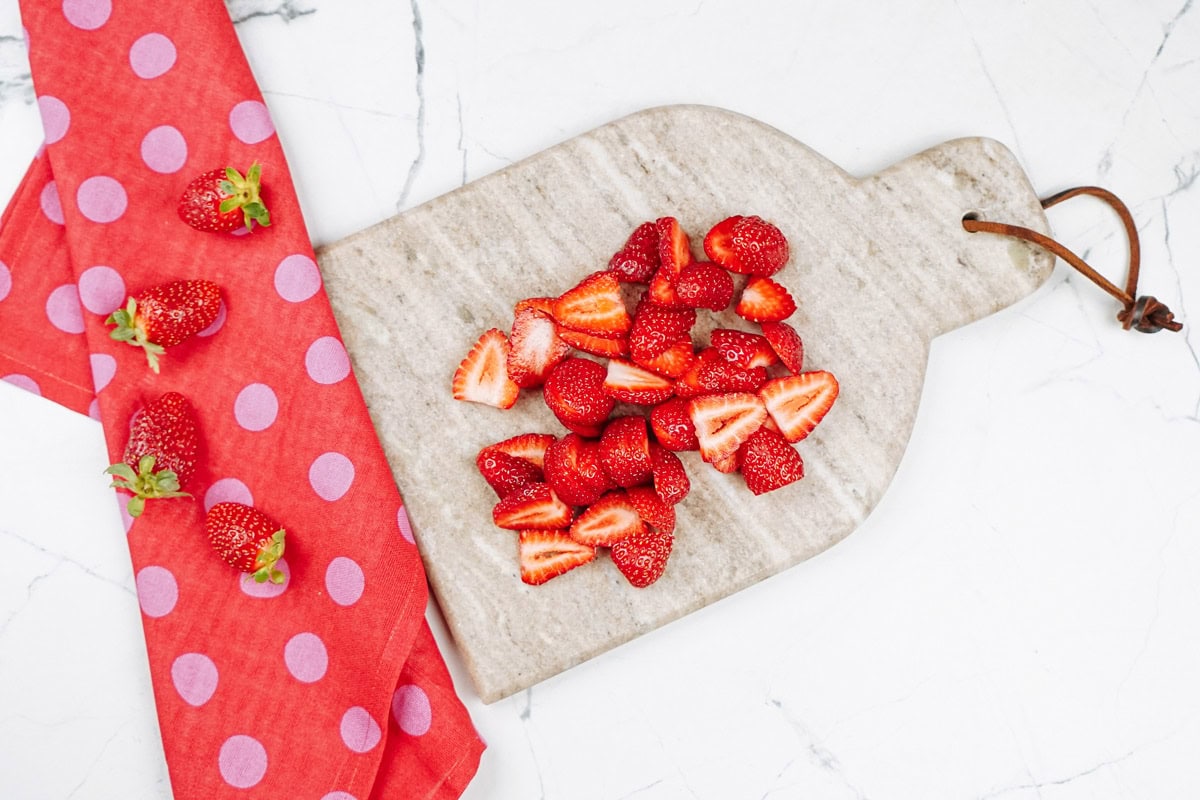 Cuted riped strawberries are on the marple cut board. Near it, there is a kitchen towel with some whole strawberries | Hurry The Food Up