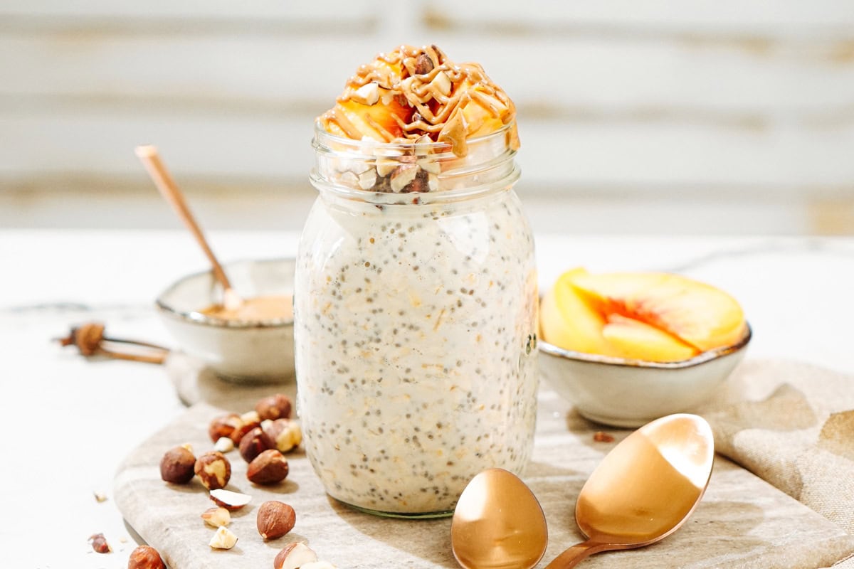 The Mason jar with overnight oats is on the marble cut board. There are some hazelnuts, two bronze spoons, a tiny bowl with peanut butter and a small dish with some slices of peach | Hurry The Food Up