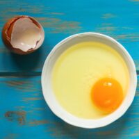 A white plate with egg yolk and white is on a blue surface. Near it, there is eggshell | Hurry The Food Up - 6