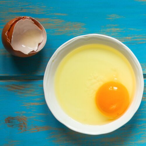 A white plate with egg yolk and white is on a blue surface. Near it, there is eggshell | Hurry The Food Up