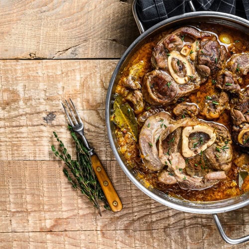 Bird's eye view of a pot with Osso buco on a wooden surface. Next to the pot is a fork, black towel, and some herbs