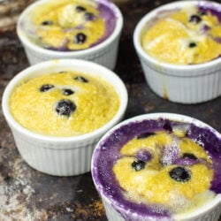 Four ramekins filled with baked blueberry desserts, with golden tops and blueberry juice visible on the surface.