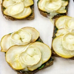 Slices of Honey Pesto Goat Cheese Toast, topped with creamy goat cheese, drizzled with golden honey, and garnished with fresh herbs, sit elegantly on a cutting board.
