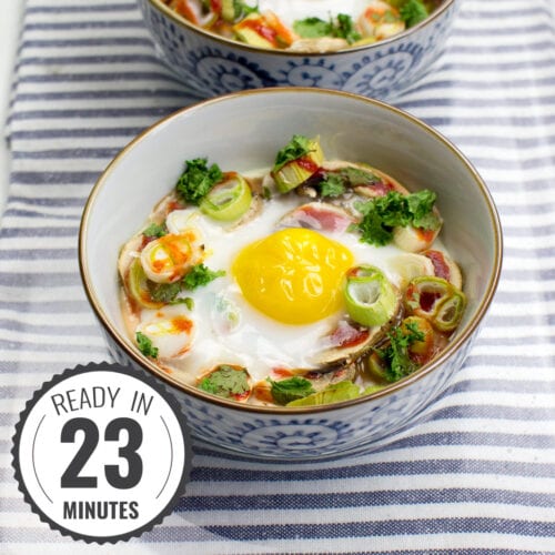 Two bowls of Mushroom Oeuf Cocotte, baked with vegetables and garnished with herbs, rest on a striped cloth inside a glass tray with a timestamp in the left corner.