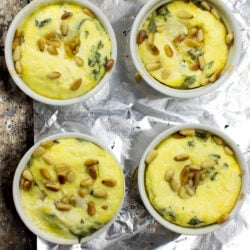 Four white ramekins filled with Caprese Quiche, featuring a yellow egg mixture with green herbs and pine nuts, placed on a sheet of aluminum foil on a textured surface.
