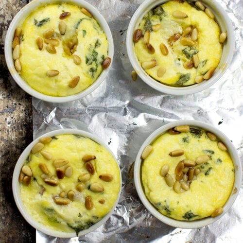 Four white ramekins filled with Caprese Quiche, featuring a yellow egg mixture with green herbs and pine nuts, placed on a sheet of aluminum foil on a textured surface.