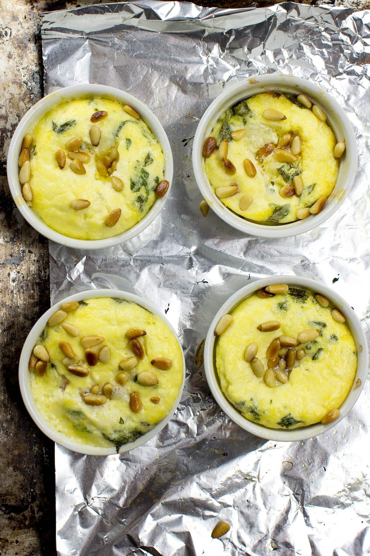 Four white ramekins filled with Caprese Quiche, featuring a yellow egg mixture with green herbs and pine nuts, placed on a sheet of aluminum foil on a textured surface.