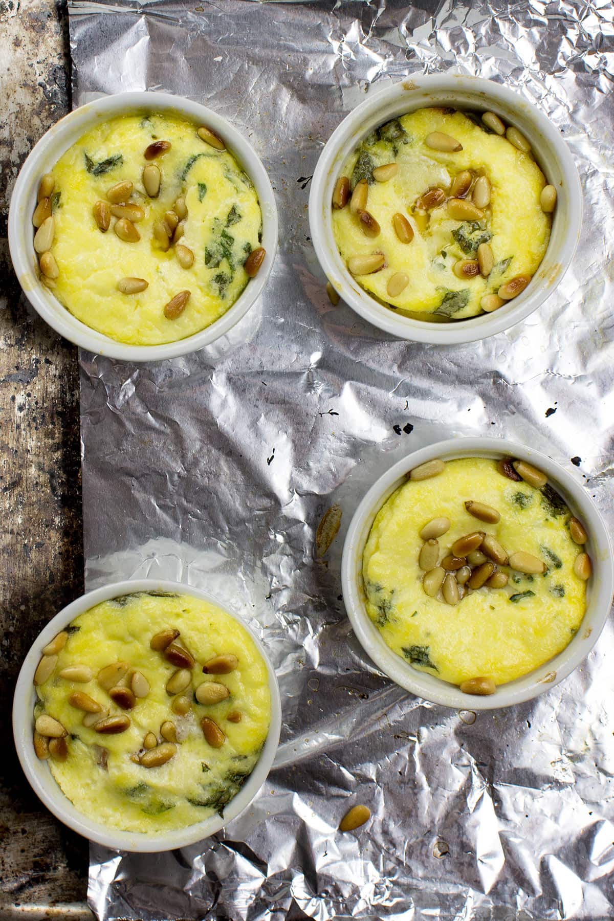 Four white ramekins filled with Caprese Quiche, featuring a yellow egg mixture with green herbs and pine nuts, placed on a sheet of aluminum foil on a textured surface.