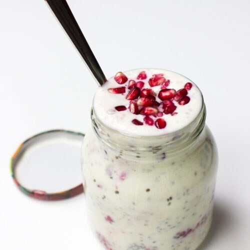 A glass jar filled with a Chia Yoghurt Bowl topped with pomegranate seeds and raspberries, with a spoon inserted and the lid placed beside it on a white surface.