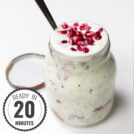 A glass jar filled with a Chia Yoghurt Bowl topped with pomegranate seeds and raspberries, with a spoon inserted and the lid placed beside it on a white surface with a timestamp in the left corner.