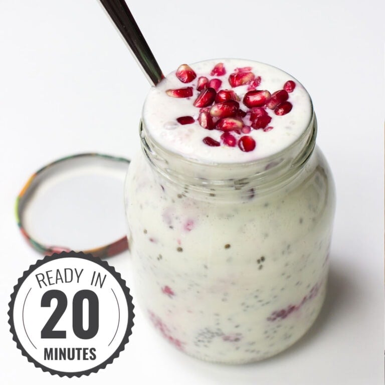 A glass jar filled with a Chia Yoghurt Bowl topped with pomegranate seeds and raspberries, with a spoon inserted and the lid placed beside it on a white surface with a timestamp in the left corner.
