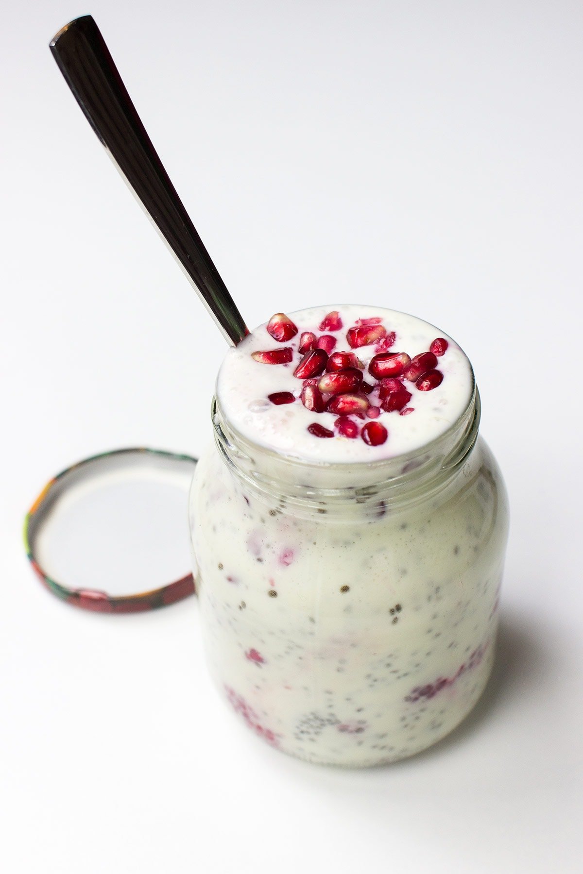 A glass jar filled with a Chia Yoghurt Bowl topped with pomegranate seeds and raspberries, with a spoon inserted and the lid placed beside it on a white surface.