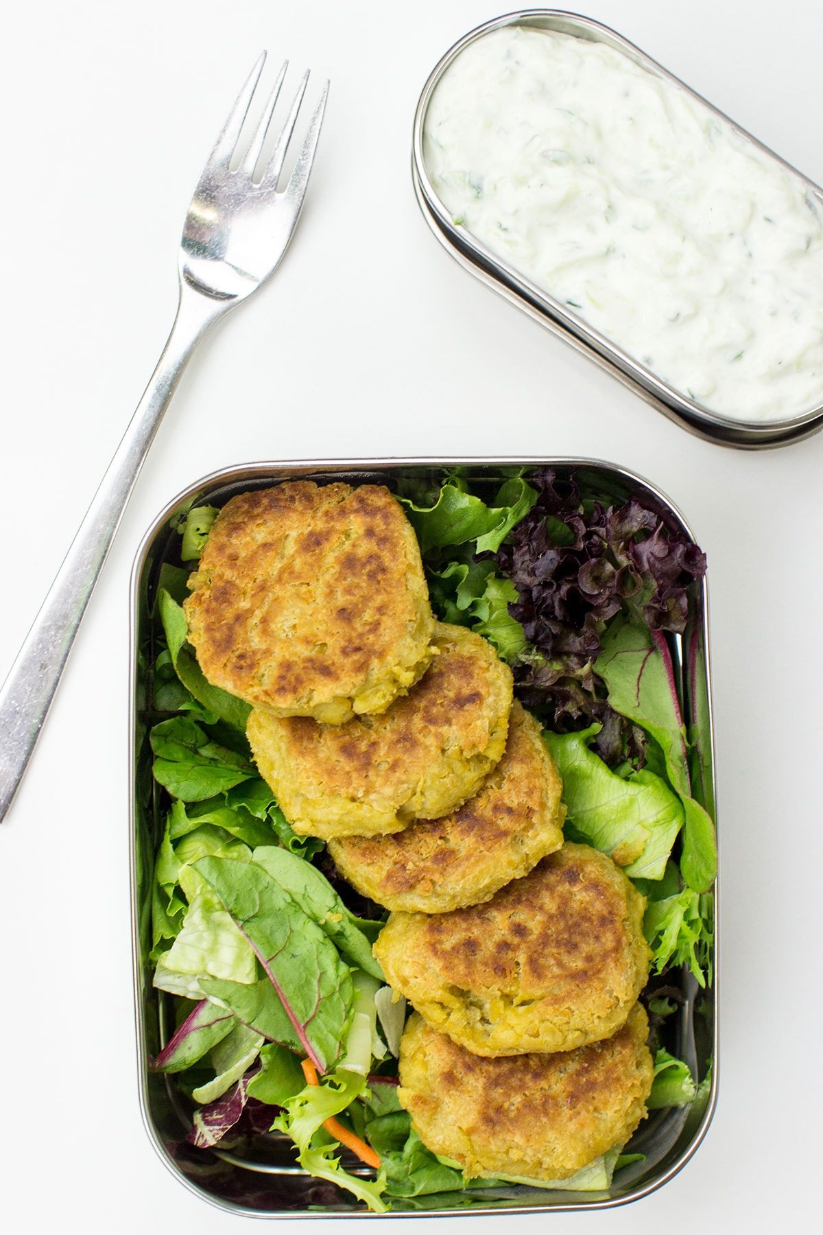 A metal lunch container with Chickpea Fritters placed on a bed of mixed greens, accompanied by a separate compartment with Cucumber Dip and a fork on a white surface.