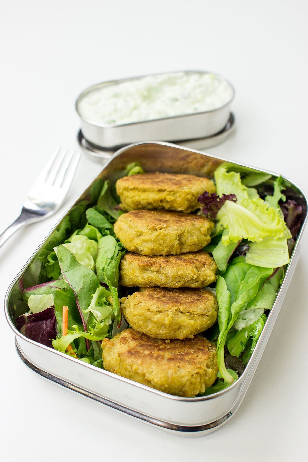 A metal lunch container with Chickpea Fritters placed on a bed of mixed greens, accompanied by a separate compartment with Cucumber Dip and a fork on a white surface.