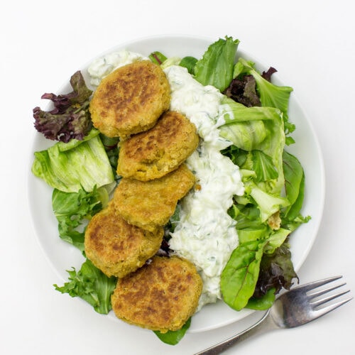A white plate with Chickpea Fritters arranged on a bed of mixed greens, topped with a generous serving of Cucumber Dip, and a fork placed beside it on a white surface.