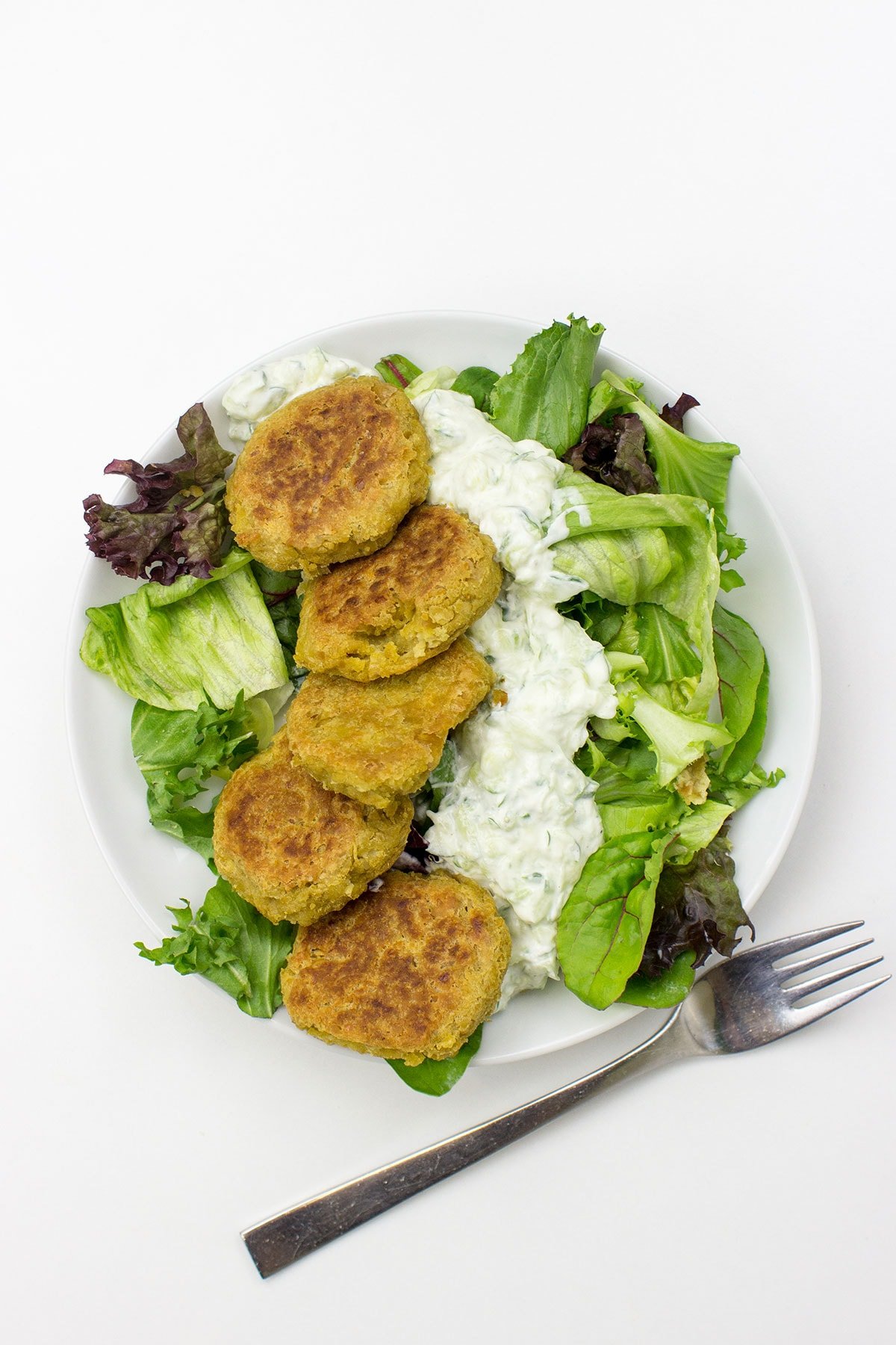 A white plate with Chickpea Fritters arranged on a bed of mixed greens, topped with a generous serving of Cucumber Dip, and a fork placed beside it on a white surface.