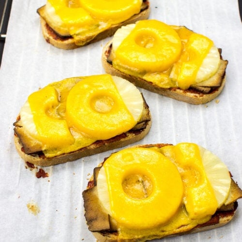 A white surface with four pieces of Hawaiian Tofu Toast topped with melted cheese and pineapple slices, arranged in a vertical line.