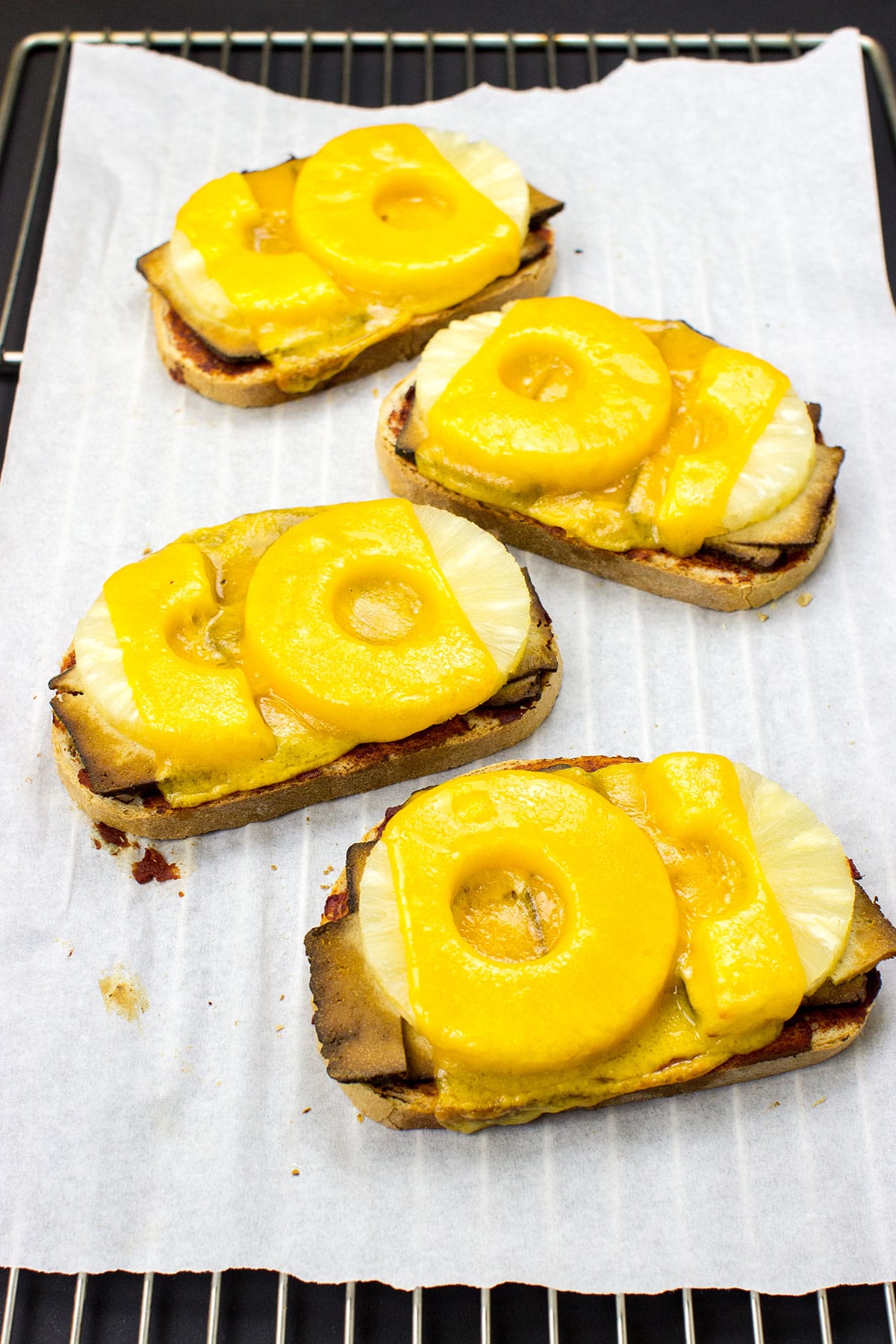 A white surface with four pieces of Hawaiian Tofu Toast topped with melted cheese and pineapple slices, arranged in a vertical line.