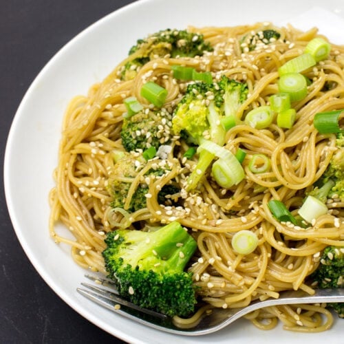A white plate topped with Hoisin Ginger Noodles, featuring broccoli, green onions, and sesame seeds, with a fork resting on the side.