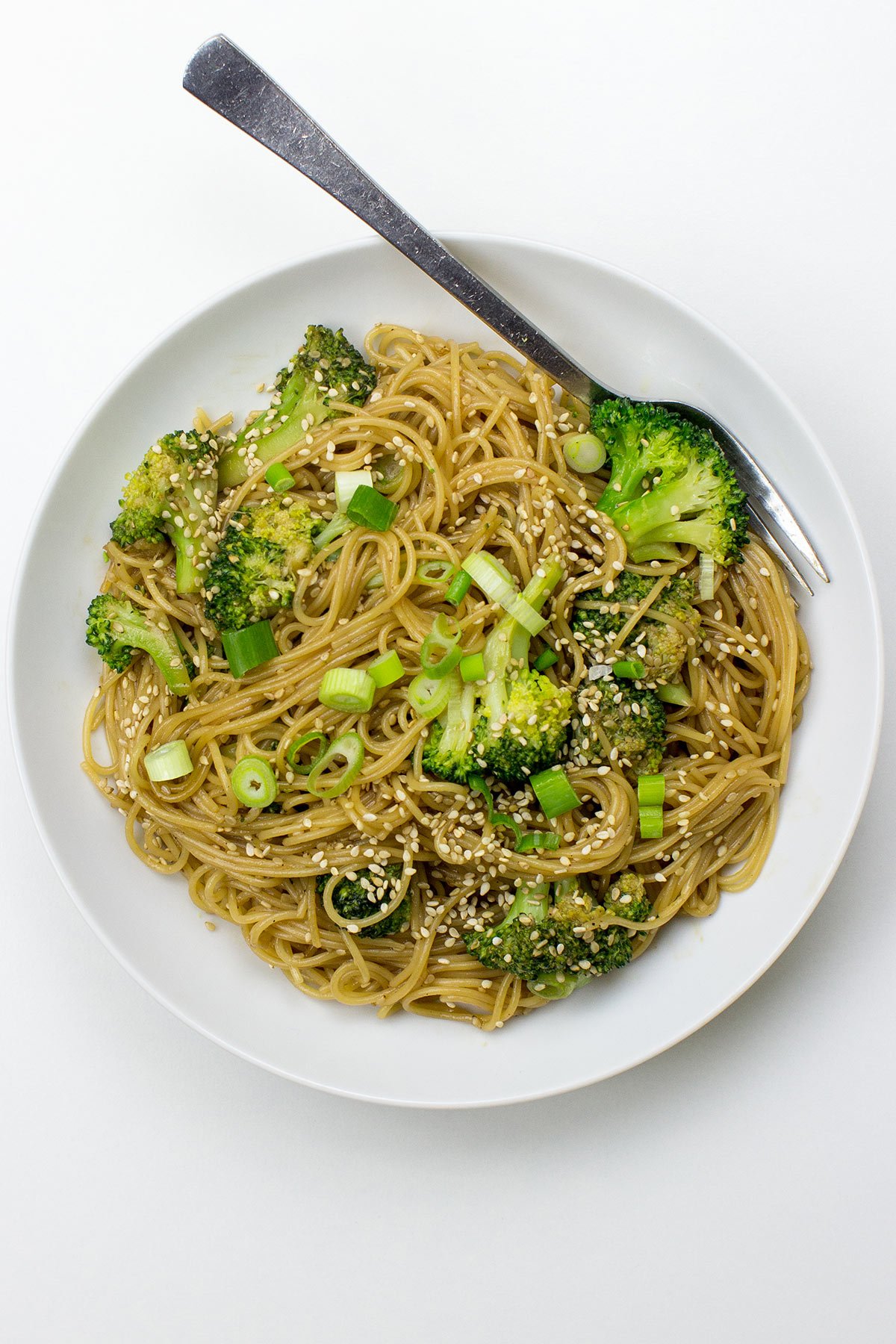 A white plate topped with Hoisin Ginger Noodles, featuring broccoli, green onions, and sesame seeds, with a fork resting on the side.