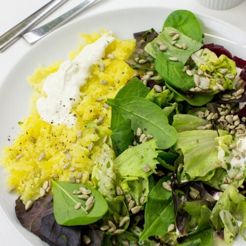 A white plate with Roasted Beetroot & Potato Salad, featuring yellow potatoes, red beetroot cubes, mixed greens, sunflower seeds, and dollops of lime crème fraîche, accompanied by a small bowl of crème fraîche, a knife, and a fork on a white surface.