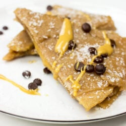 A white plate with two Vegan Chocolate & Coconut Crepes, topped with shredded coconut, chocolate chips, and drizzled with yellow sauce.