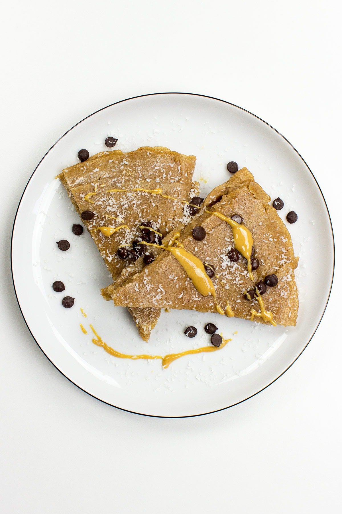 A white plate with two Vegan Chocolate & Coconut Crepes, topped with shredded coconut, chocolate chips, and drizzled with yellow sauce.